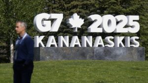 A man stands in front of a large white sign reading “G7 2025 Kananaskis” with a maple leaf symbol, located on a grassy lawn with green pine trees in the background.
