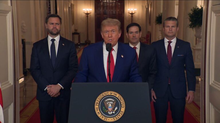 Donald Trump speaks at a podium with the Seal of the President of the United States, flanked by three suited men, delivering a serious statement in a formal hallway.